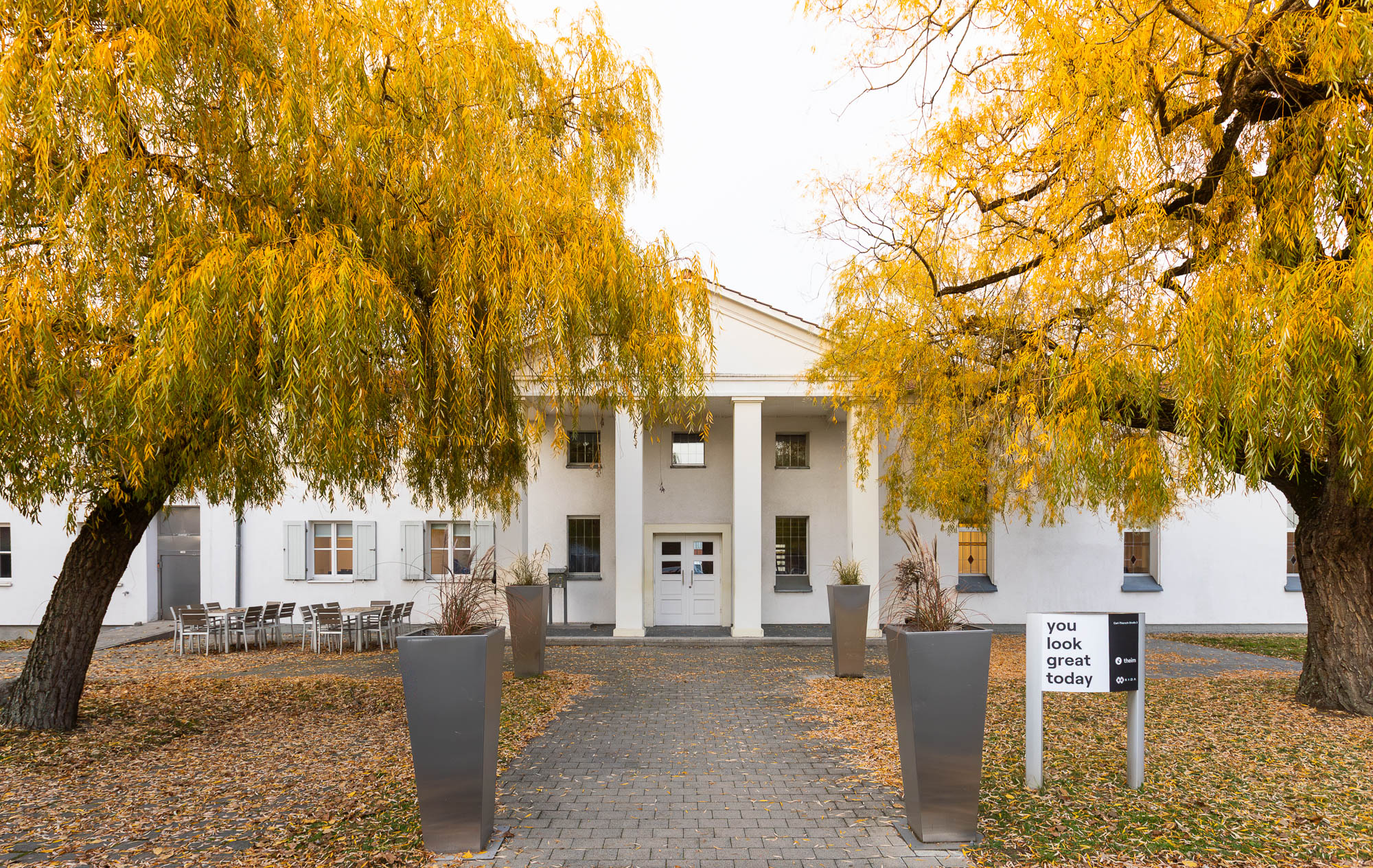 Weißes Agenturgebäude mit Säulen und herbstlichen Bäumen im Innenhof.